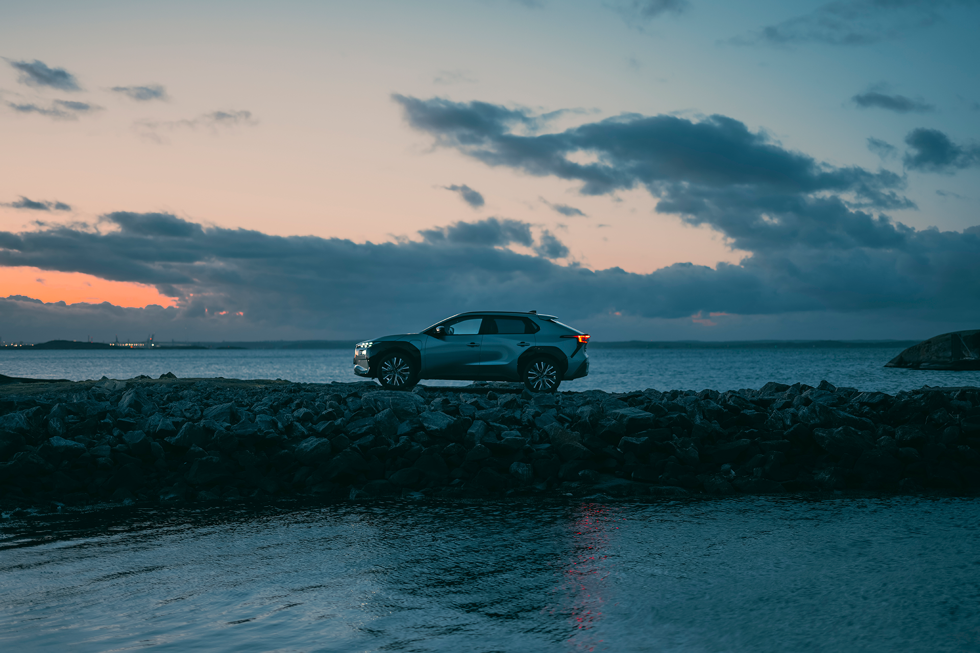 Subaru Solterra novietots akmeņainā jūras piekrastē saulrieta laikā.